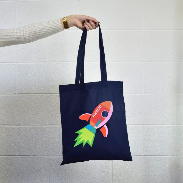 hand holding a navy blue tote bag with pink Monzo cartoon space rocket