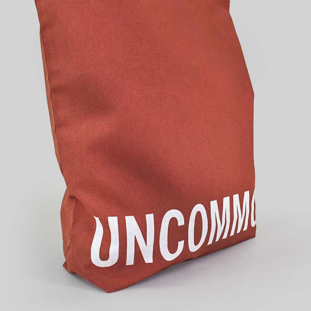 rust red tote bag with white text that says UNCOMMON on bottom panel of bag