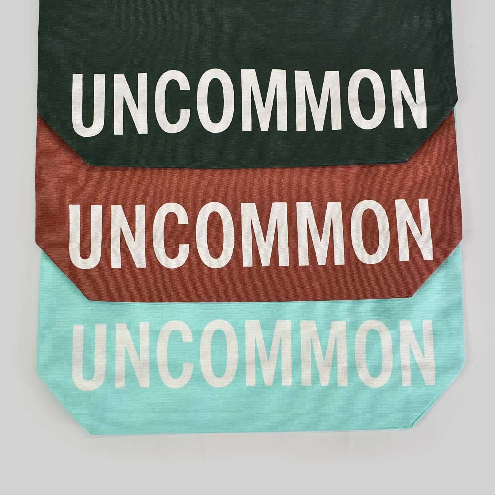 Three bags shown one in turquoise, one in rust red and the other in dark green both have white handles and white text printed along the bottom panel of the bag that says UNCOMMON