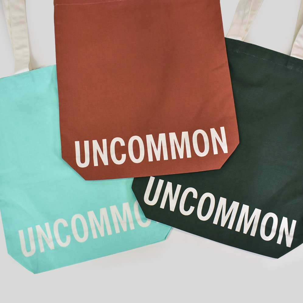 Three bags shown one in turquoise, one in rust red and the other in dark green both have white handles and white text printed along the bottom panel of the bag that says UNCOMMON